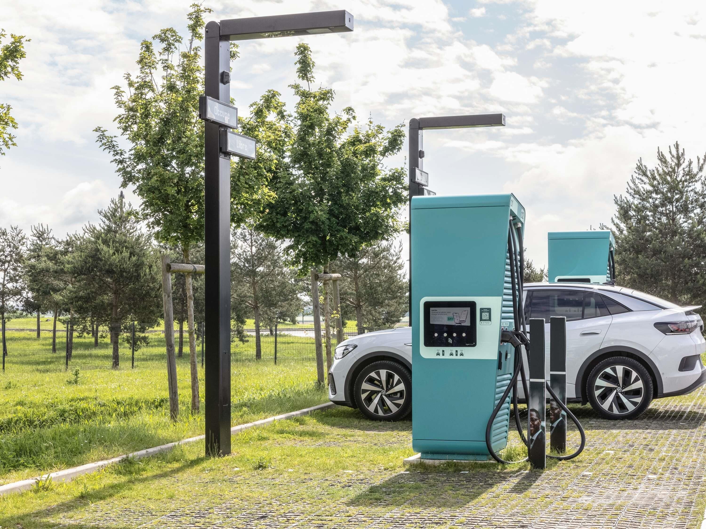 A white car parked at a charging station with a teal charging unit, surrounded by greenery and trees.