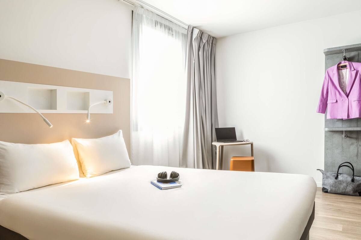 A neatly made bed with white linens, a laptop on a small table beside it, and a pink blazer hanging on a hook.