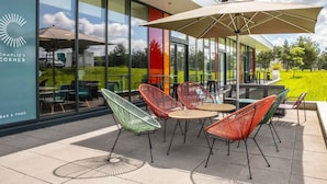 An outdoor seating area with tables and chairs, a large umbrella, and a view of a building with a glass facade. - ibis budget Gonesse Le Bourget (Gonesse)