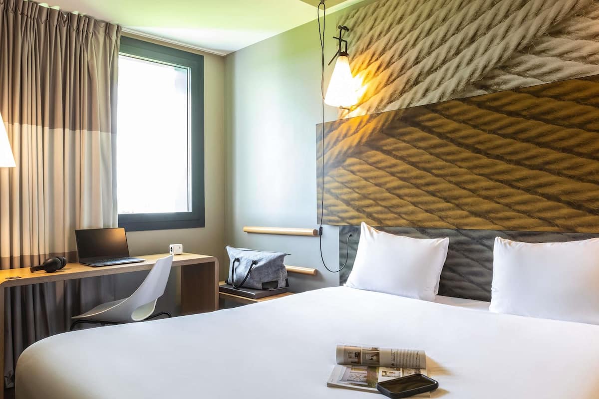 A hotel room with a large bed, a desk with a laptop, a chair, a lamp, and a window with curtains.