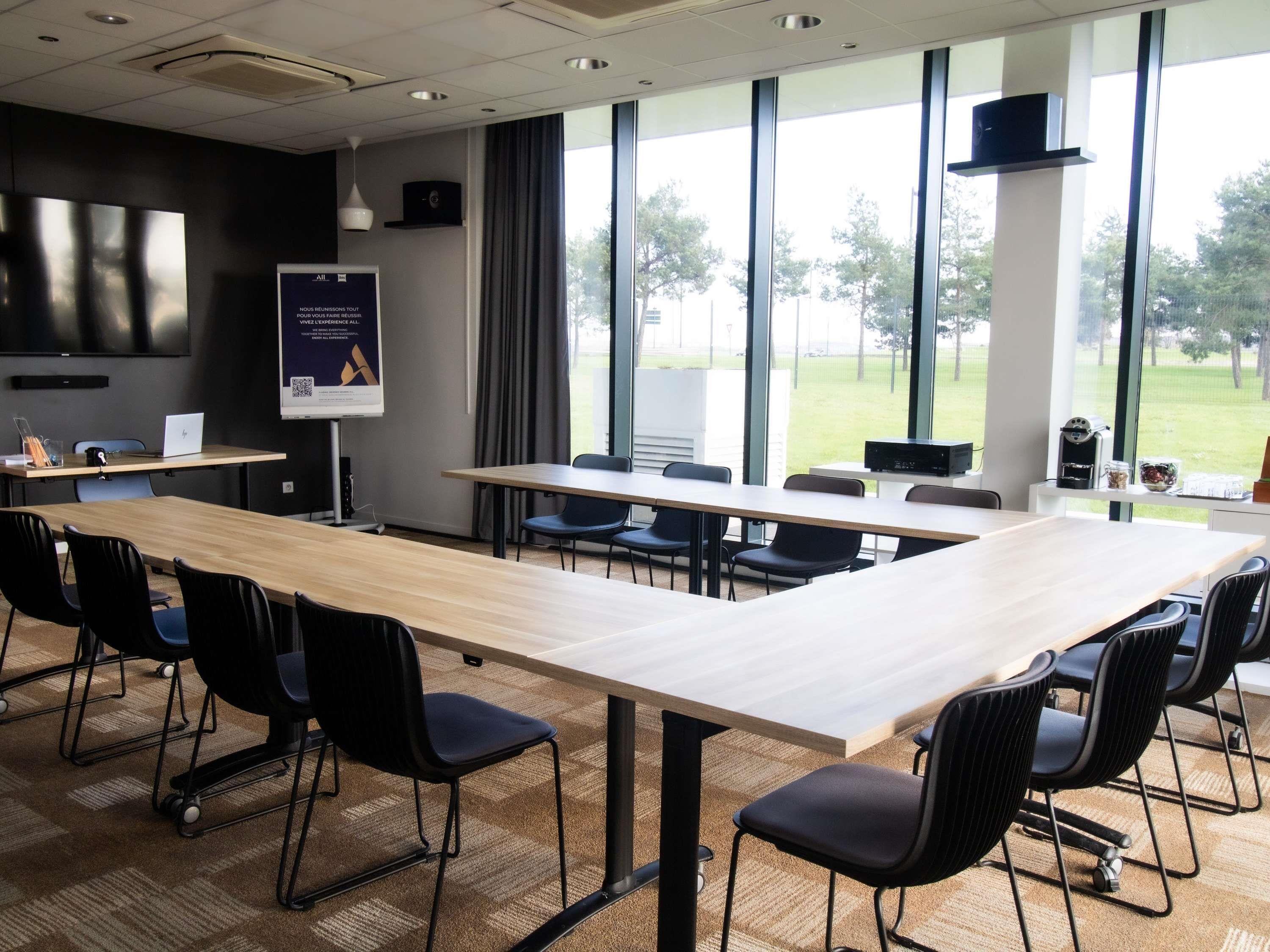A conference room with a long table, black chairs, a projector screen, and a window with a view of trees.