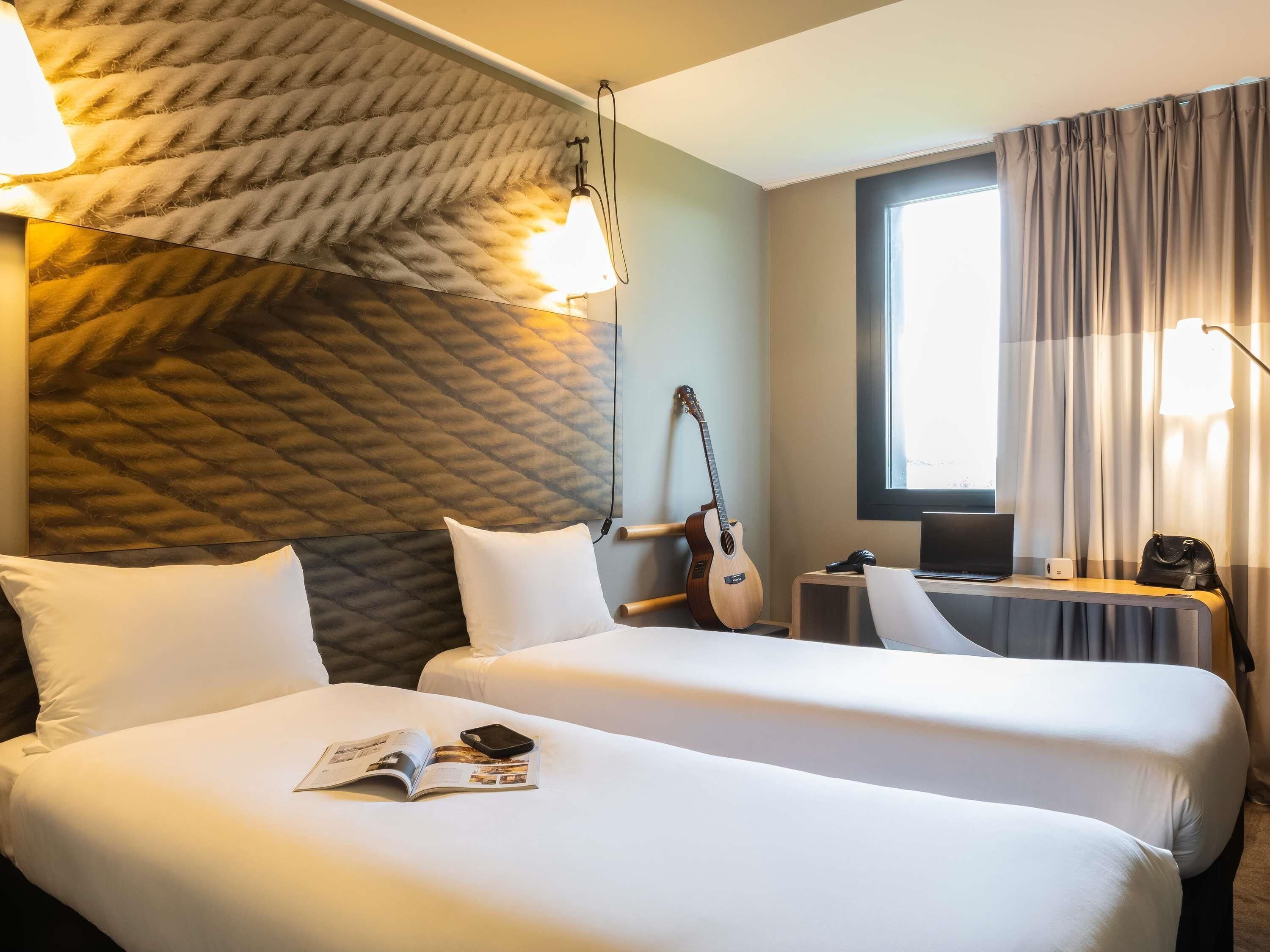 Une chambre d’hôtel avec deux lits, une guitare, un ordinateur portable et une fenêtre avec des rideaux.
