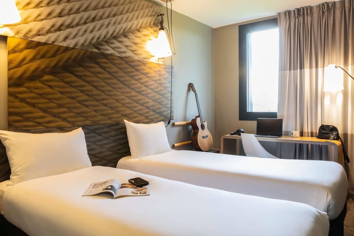 A hotel room with two beds, a guitar, a laptop, and a window with curtains.