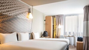 Habitación de hotel con dos camas, un escritorio, una silla, una lámpara y un ventanal con cortinas.