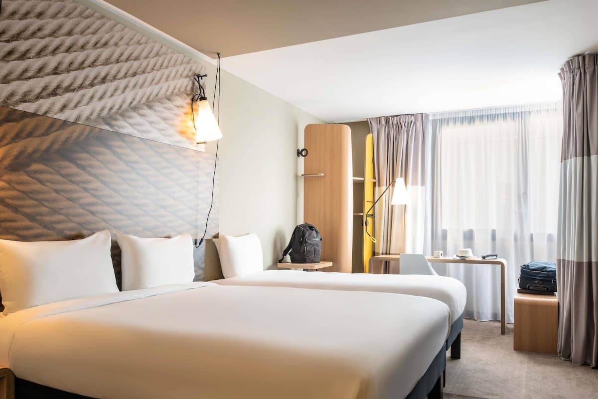 A hotel room with two beds, a desk, a chair, a lamp, and a window with curtains.