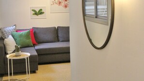 Ett vardagsrum med en grå soffa, ett litet vitt sidobord, en spegel och ett fönster med persienner.