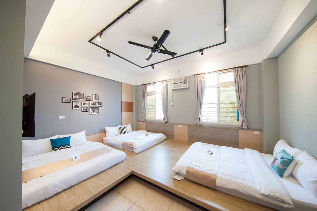 A hotel room with three beds, a ceiling fan, an air conditioning unit, and a window with curtains.