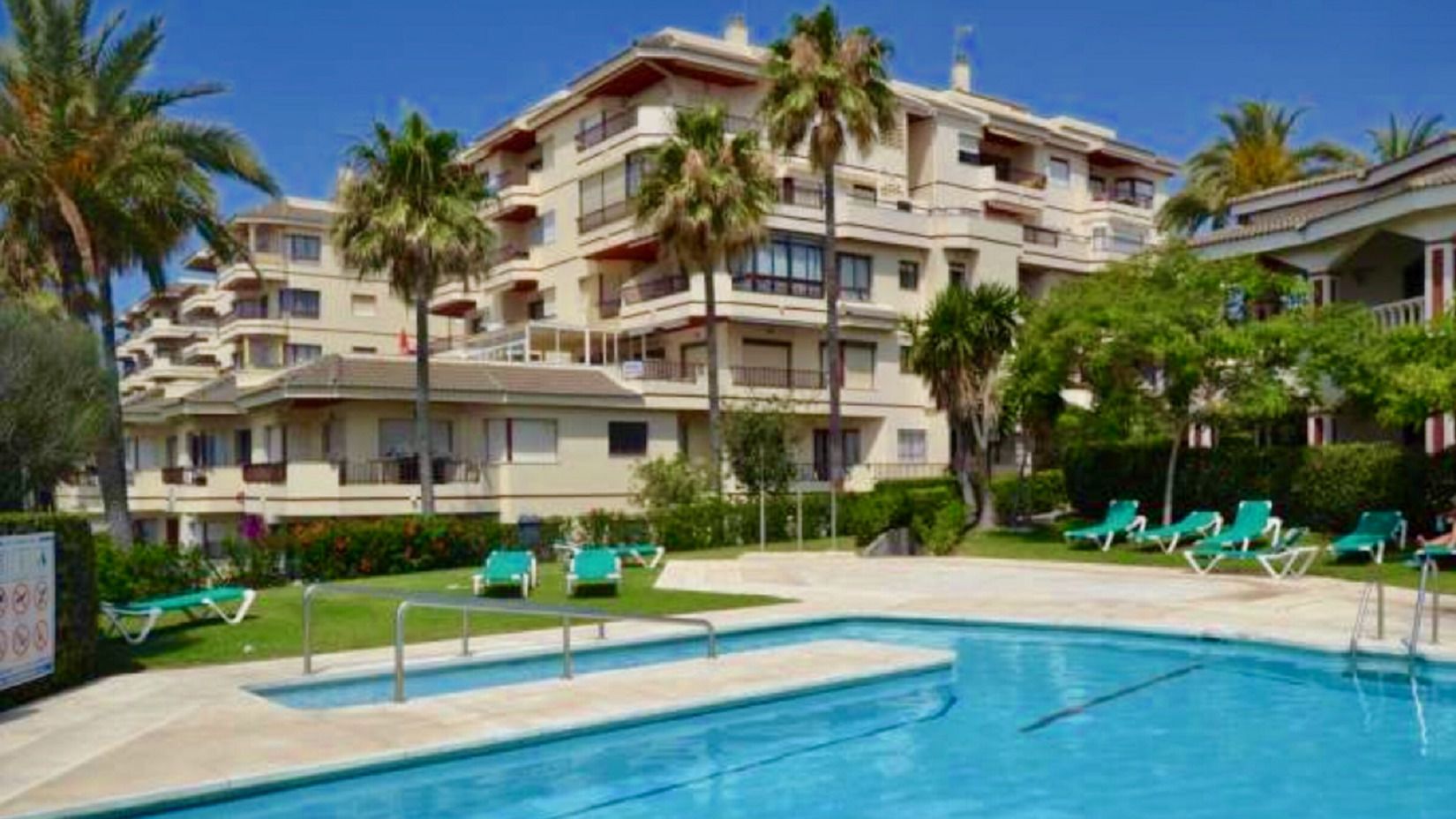 A hotel with a swimming pool, green lounge chairs, and palm trees.