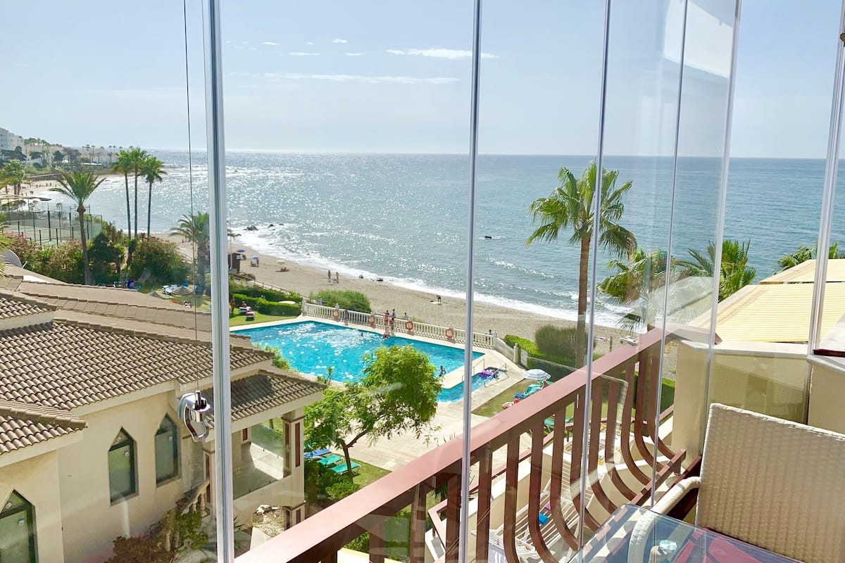 Vista desde un balcón sobre una playa, una piscina y un cielo azul despejado.