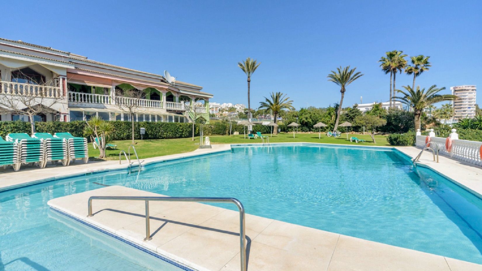 A swimming pool with a clear blue water, surrounded by a white deck and a few palm trees.