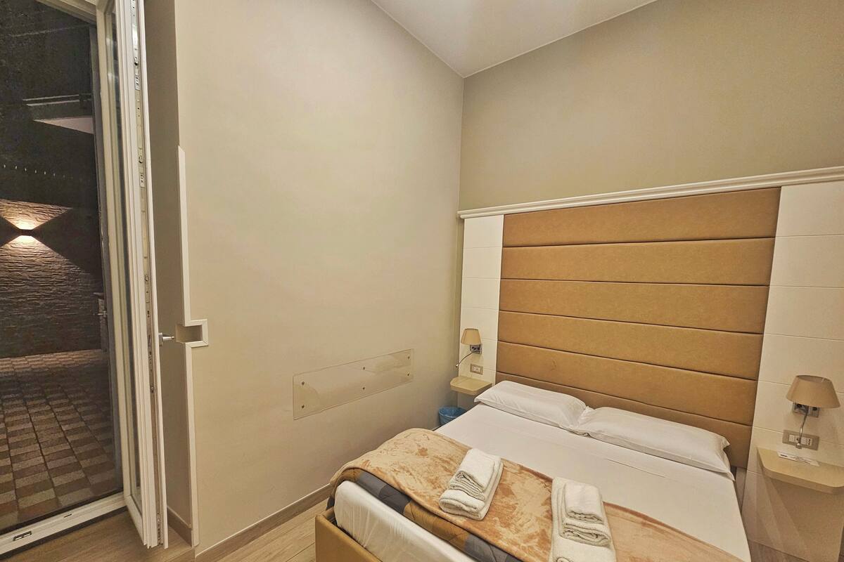 Ein Hotelzimmer mit Bett, Nachttisch, Wandlampe und Blick ins Freie.