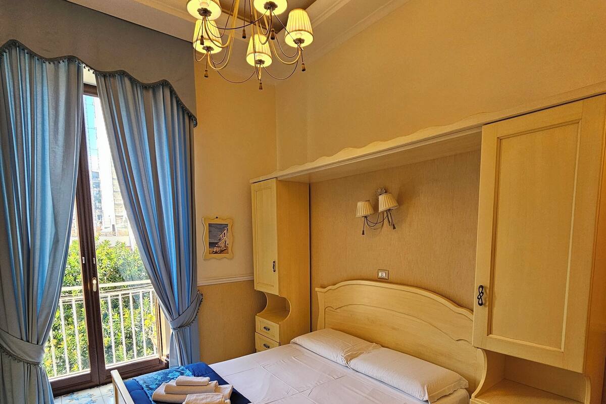 Ein Hotelzimmer mit Bett, Nachttisch, Kleiderschrank, einer Kronleuchter und einem Balkon mit Vorhängen.