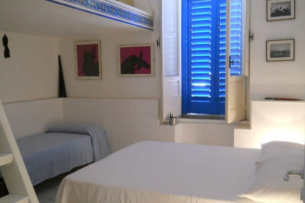 Une chambre à coucher avec un lit, un bureau et une fenêtre avec des volets bleus. Deux tableaux encadrés sont accrochés au mur.