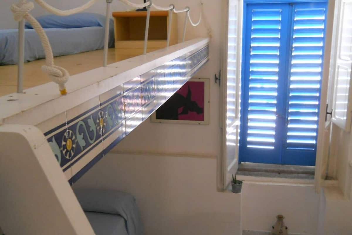 Une chambre avec un lit superposé, une petite fenêtre avec des volets bleus et un sol carrelé.