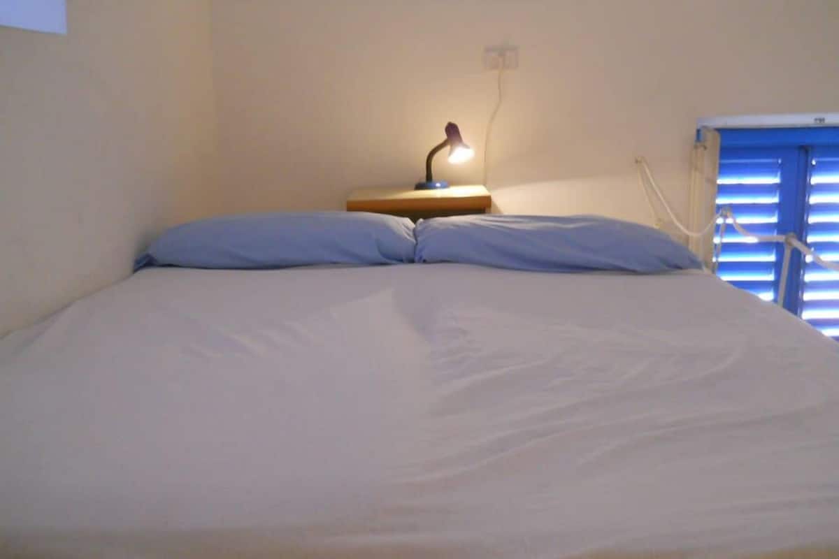 Un lit avec une table de chevet en bois et une lampe, une fenêtre avec des stores et une prise électrique au mur.