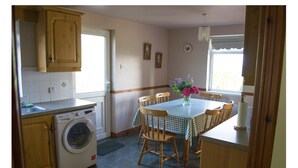 A compact kitchen with a washing machine, wooden cabinets, a dining table with chairs, and a window with curtains. - Beachview Cottage (Ballymena)