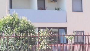 Um edifício com vários andares, varandas, uma cerca vermelha e vegetação em primeiro plano.