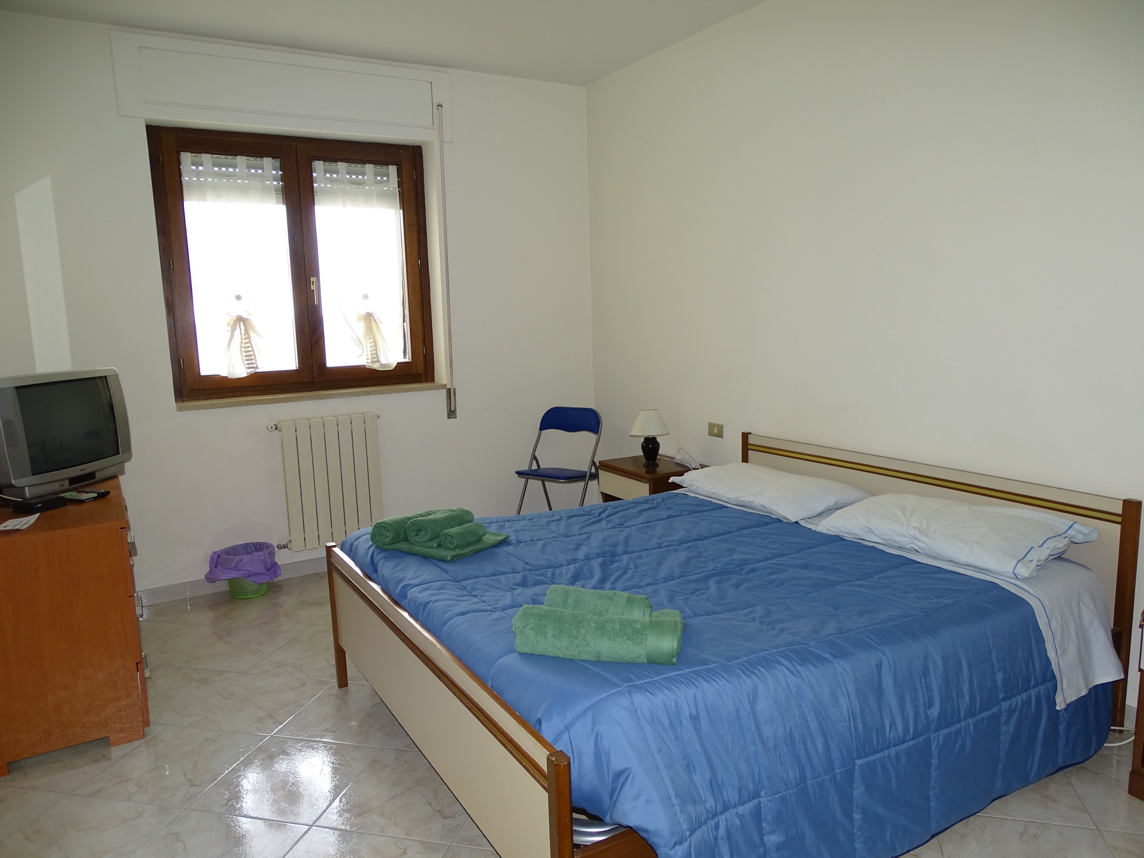 A bedroom with a bed, a television on a wooden stand, a window with curtains, a chair, and a nightstand with a lamp.