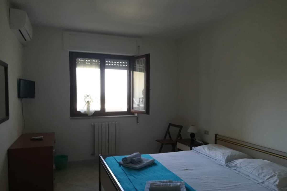 Une chambre à coucher avec un lit, une petite table, une chaise, un radiateur et une fenêtre avec des rideaux.