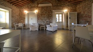 A spacious room with a high ceiling, stone walls, and a tiled floor. There are white chairs and a table, and a bookshelf is visible in the background. - Ostello Palazzo Nizza (Massa)