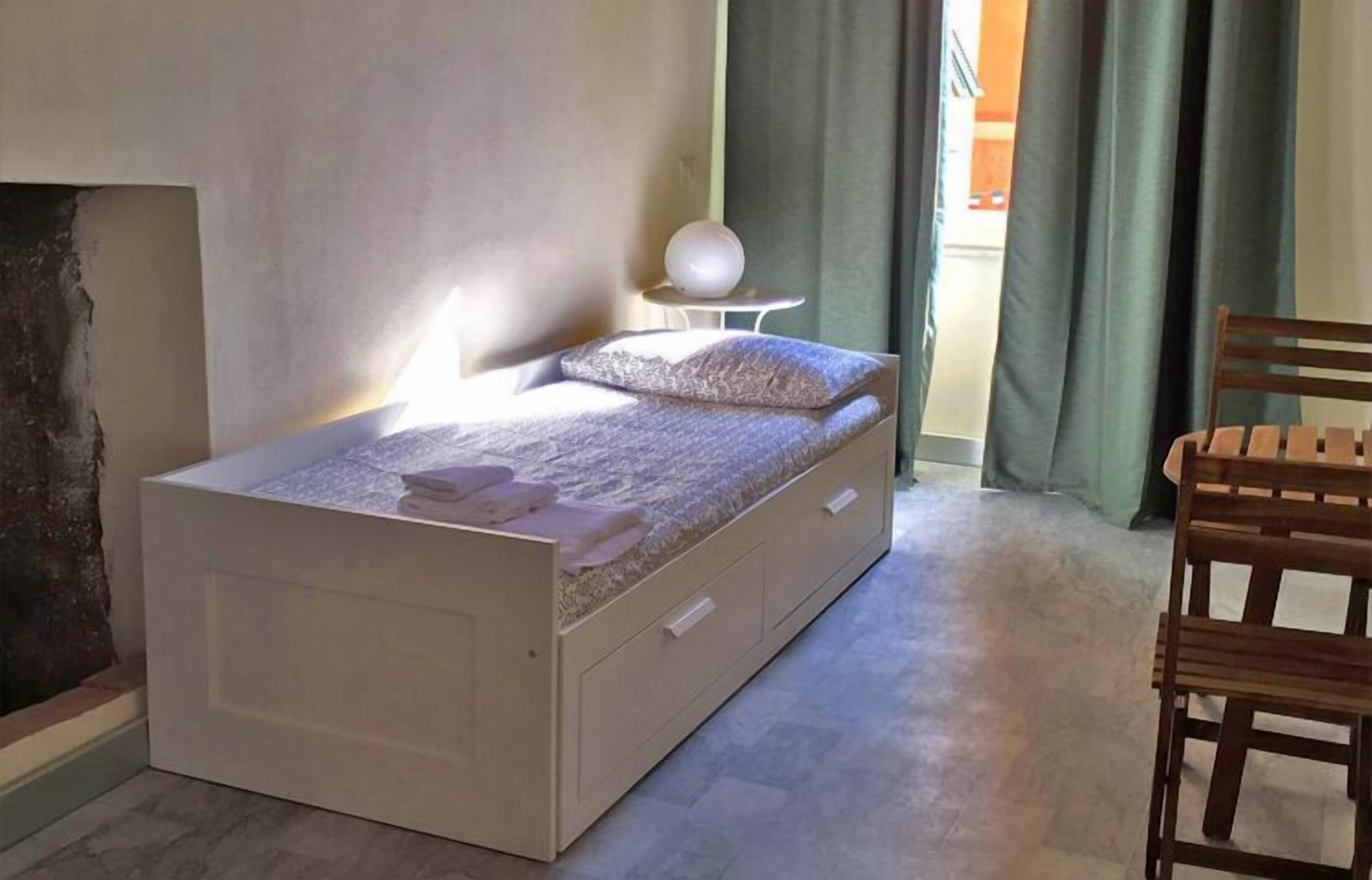 Una cama individual con un cajón para guardar objetos, una mesita de noche con una lámpara esférica y una silla.