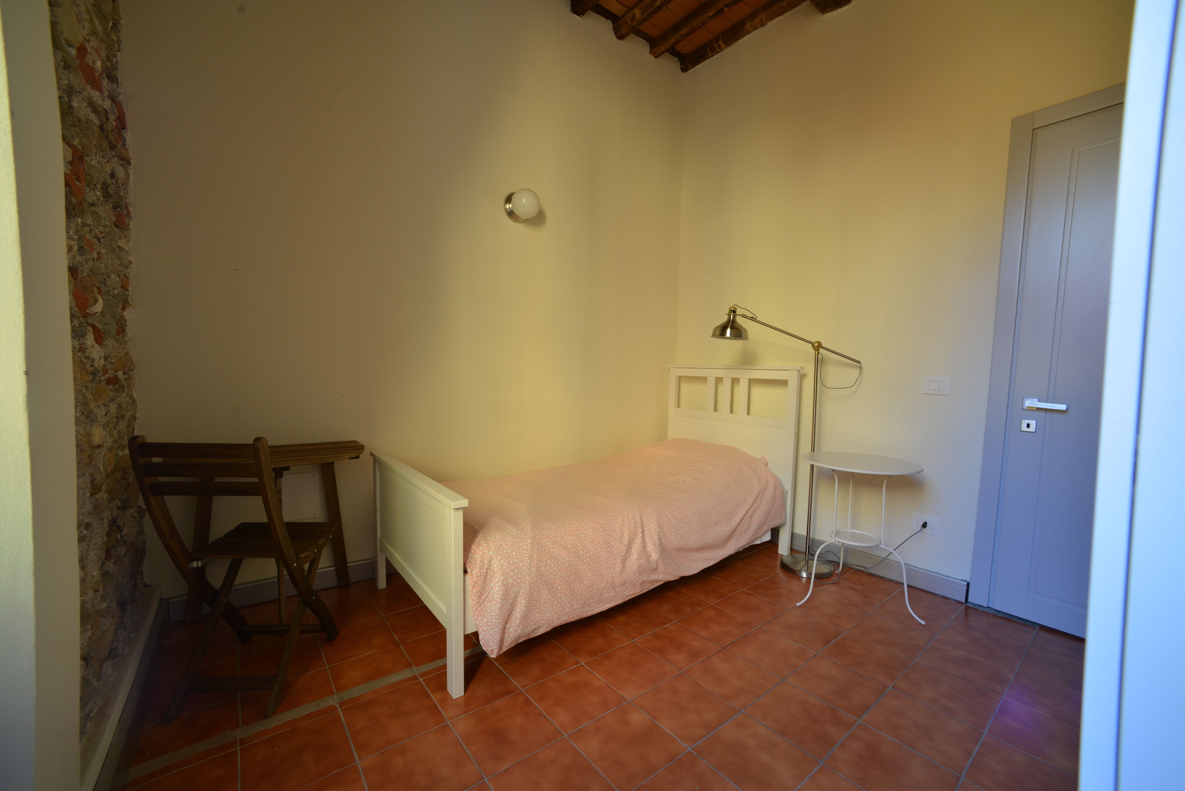 A small, single-bed room with a wooden chair, a small table, and a lamp.