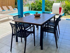 Un espace aménagé au bord de la piscine, avec une table en bois, des chaises noires et un panier en osier rempli de fruits.