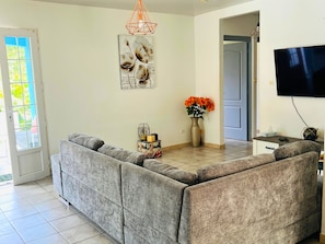 Un salon comprenant un canapé d’angle, un téléviseur à écran plat, un élément décoratif mural et un vase avec des fleurs.