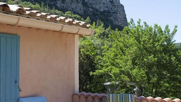 A small outdoor seating area with two chairs and a table, a building with a blue door, and a rocky cliff in the background. - Auberge De La Vallée (Saint-Jean-de-Bueges)