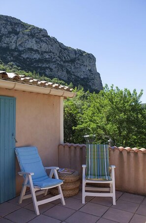 Un pequeño espacio al aire libre con dos sillas y una mesa, un edificio con puerta azul, y un risco rocoso al fondo.