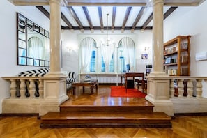 Amplia sala de estar con piso de madera, chimenea, un ventanal con cortinas, una estantería y una escalera que sube a otro nivel.
