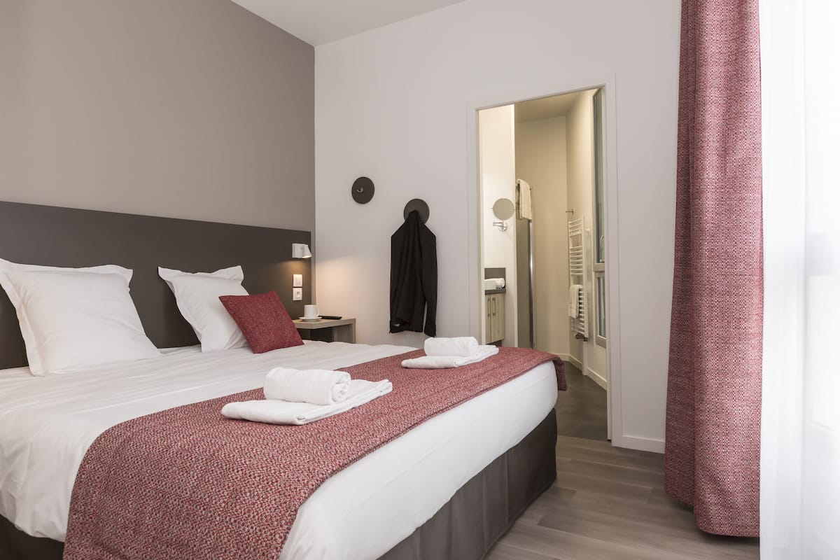 A hotel room with a large bed, a bedside table with a lamp, a mirror, and a door leading to a bathroom.