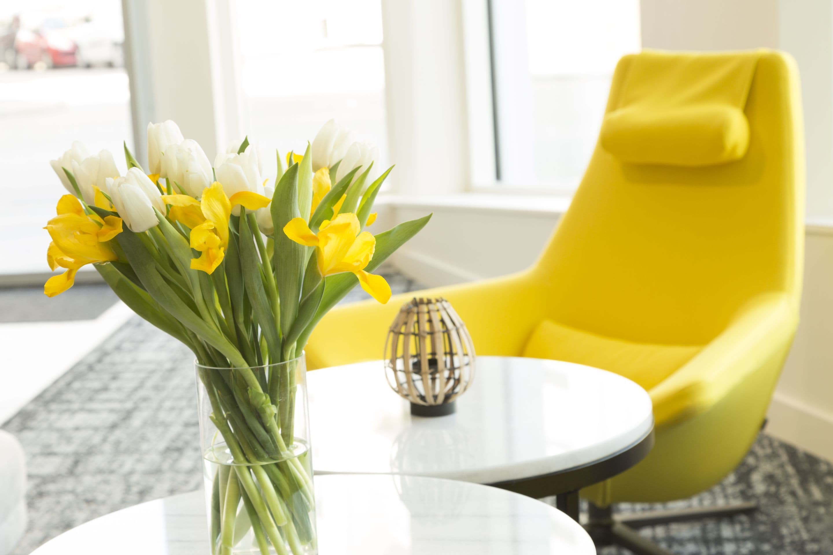 A room with a round table, a vase with flowers, a yellow chair, and a decorative lamp.