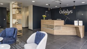 A reception area with a wooden reception desk, a white sofa with a blue pillow, and a blue armchair. - Appart'hôtel Odalys City - Le Mans Centre Palais des Congrès (Le Mans)