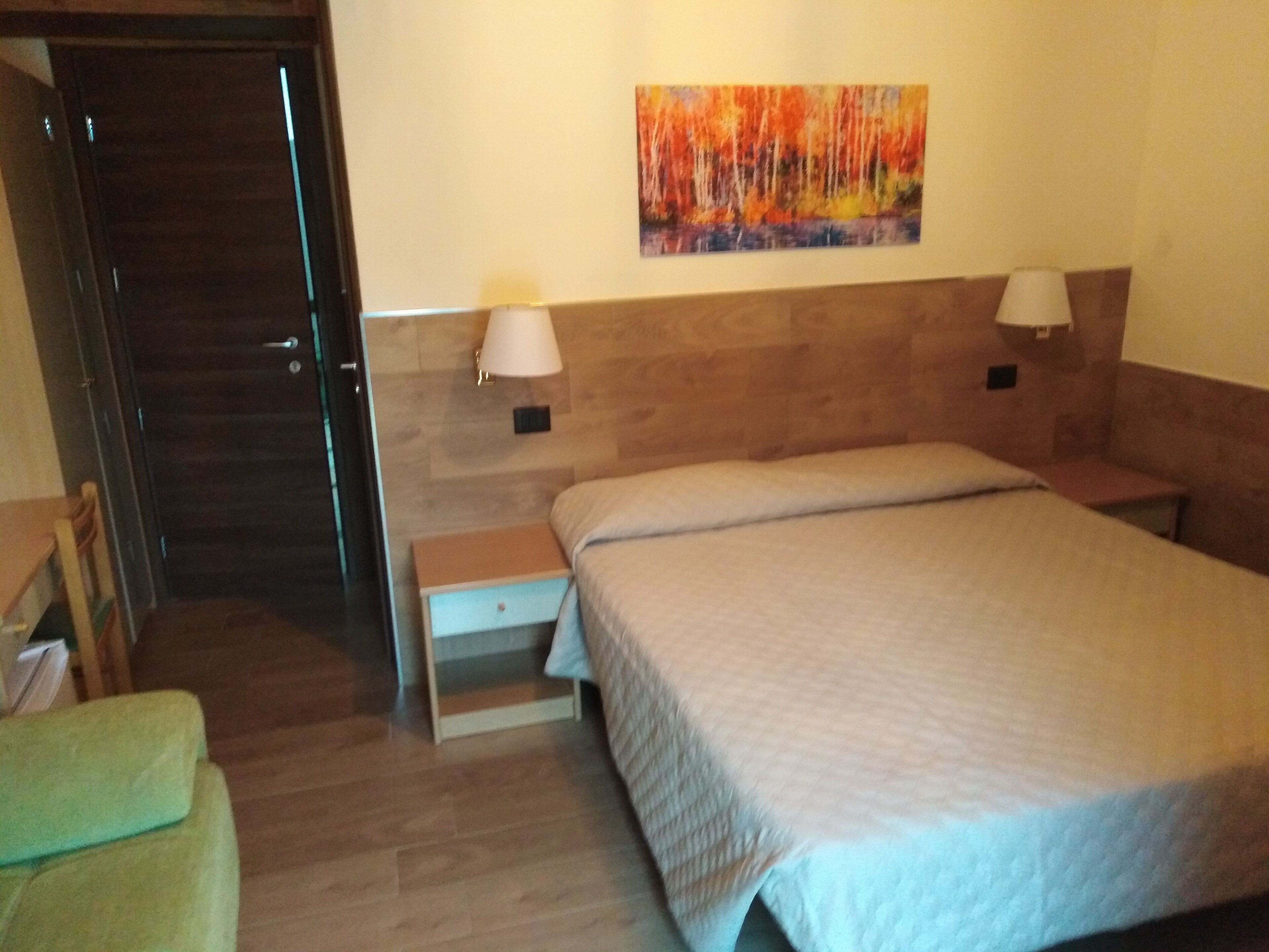 A hotel room with a bed, a nightstand, a lamp, a painting on the wall, and a wooden door.