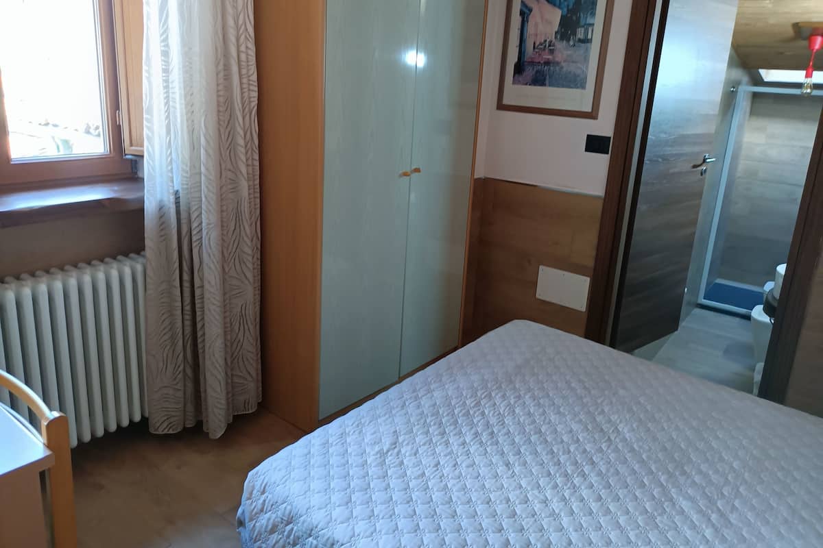 A bedroom with a bed, a wardrobe, a radiator, a window with curtains, and a door leading to a bathroom.