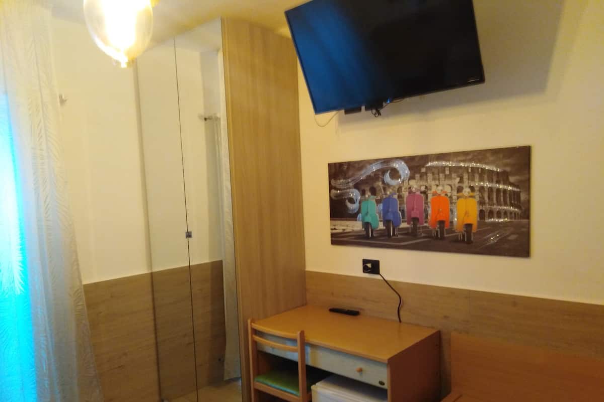 A hotel room with a bed, a desk, a small refrigerator, a wardrobe, a television mounted on the wall, and a painting of people in colorful coats.