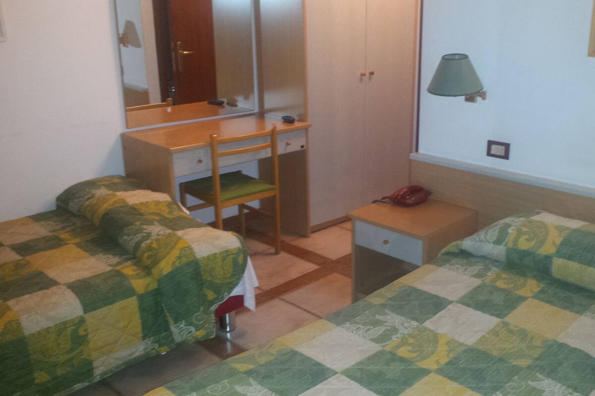 A small, neatly arranged bedroom with two single beds, a wooden desk, a chair, a wardrobe, and a bedside lamp.