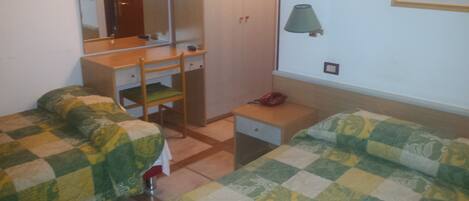Un dormitorio pequeño y ordenado con dos camas individuales, un escritorio de madera, una silla, un armario y una lámpara de noche.