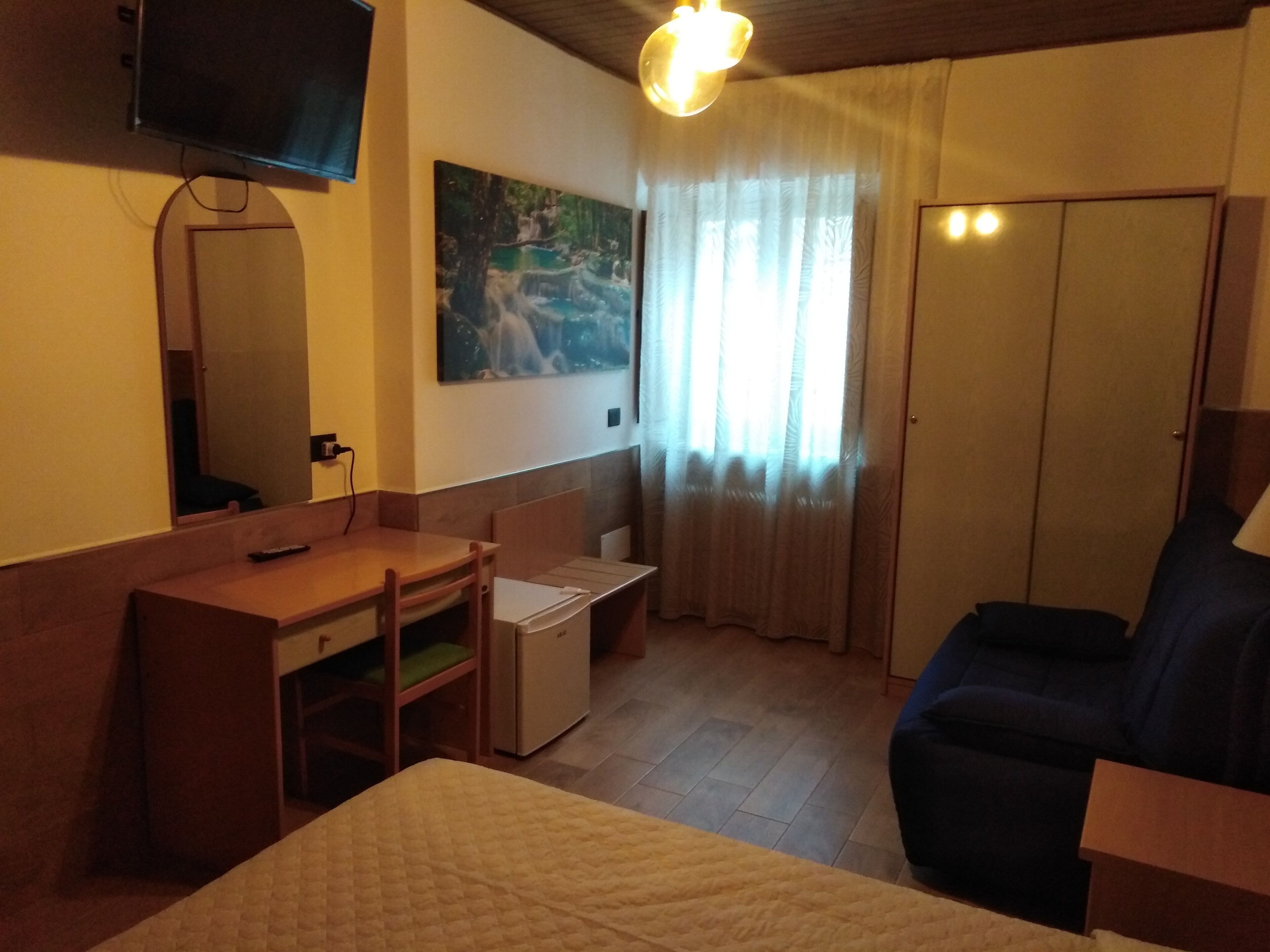 A hotel room with a bed, a desk, a chair, a small refrigerator, a television mounted on the wall, and a window with curtains.