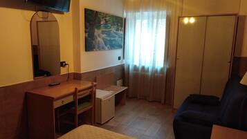 A hotel room with a bed, a desk, a chair, a small refrigerator, a television mounted on the wall, and a window with curtains.
