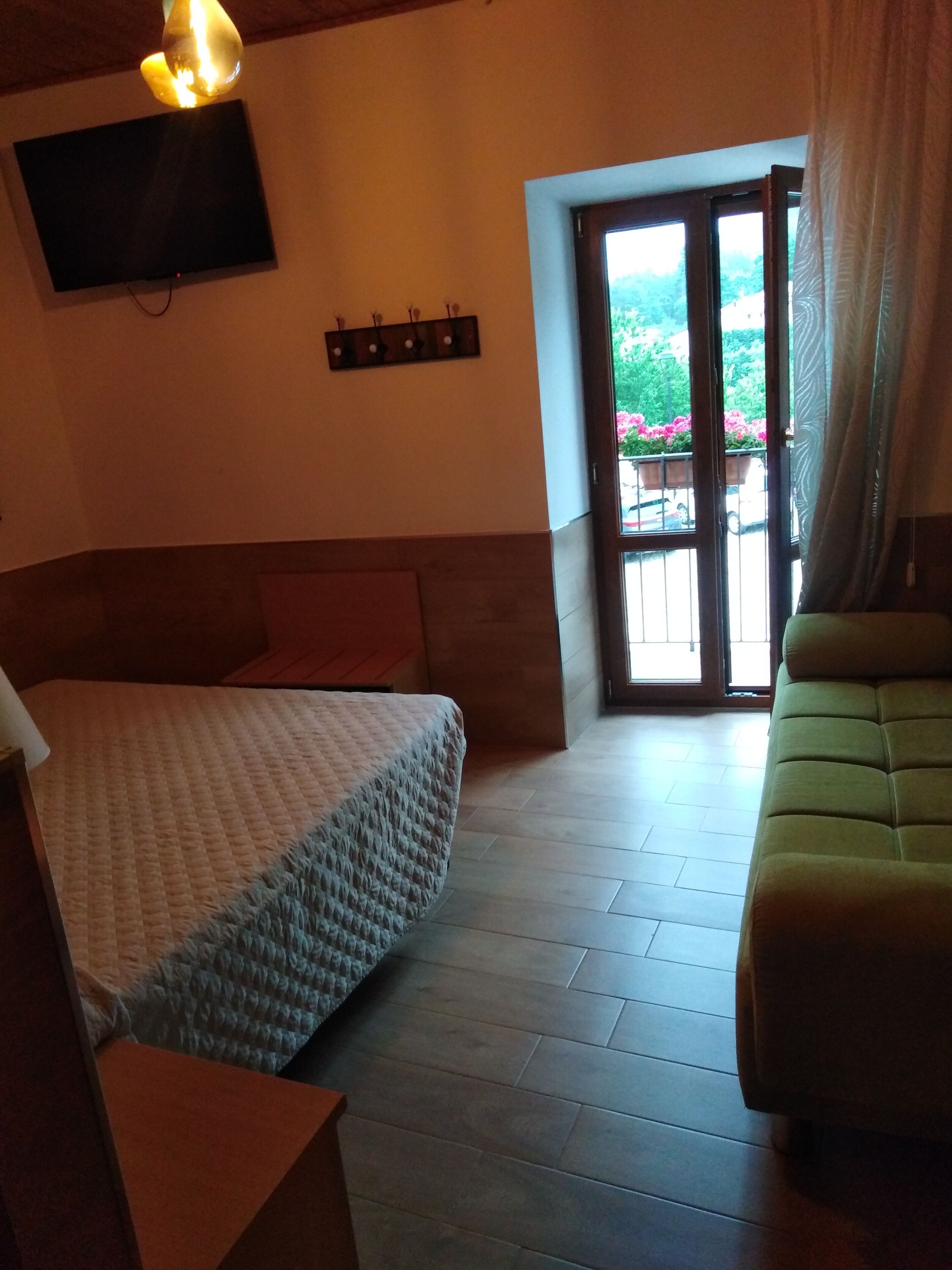 A room with a bed, a sofa, a TV mounted on the wall, and a window with a view of the outside.