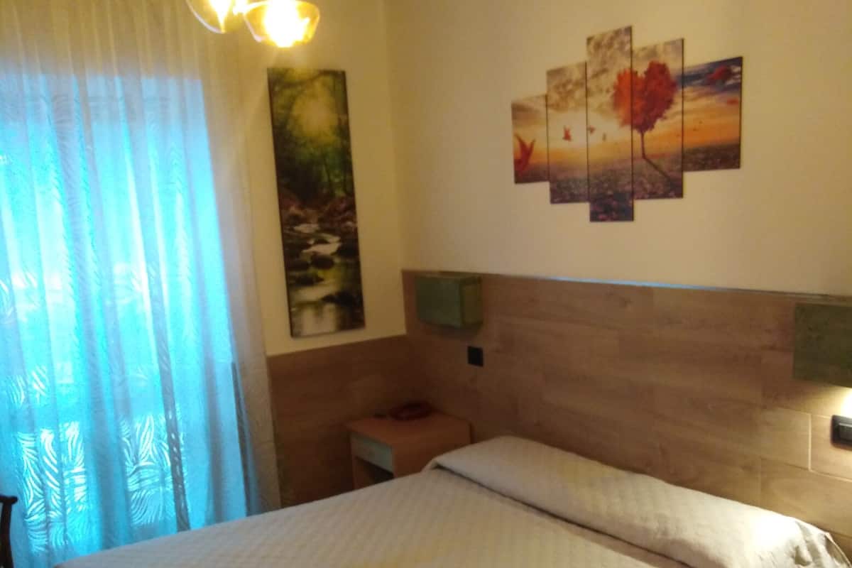 A bedroom with a wooden headboard, a bed with white bedding, a nightstand, a painting on the wall, and a window with sheer curtains.