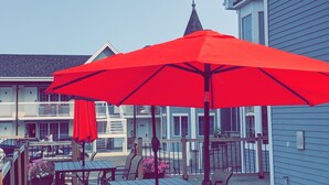 An outdoor patio with wooden tables and benches, red umbrellas, and a building in the background.
