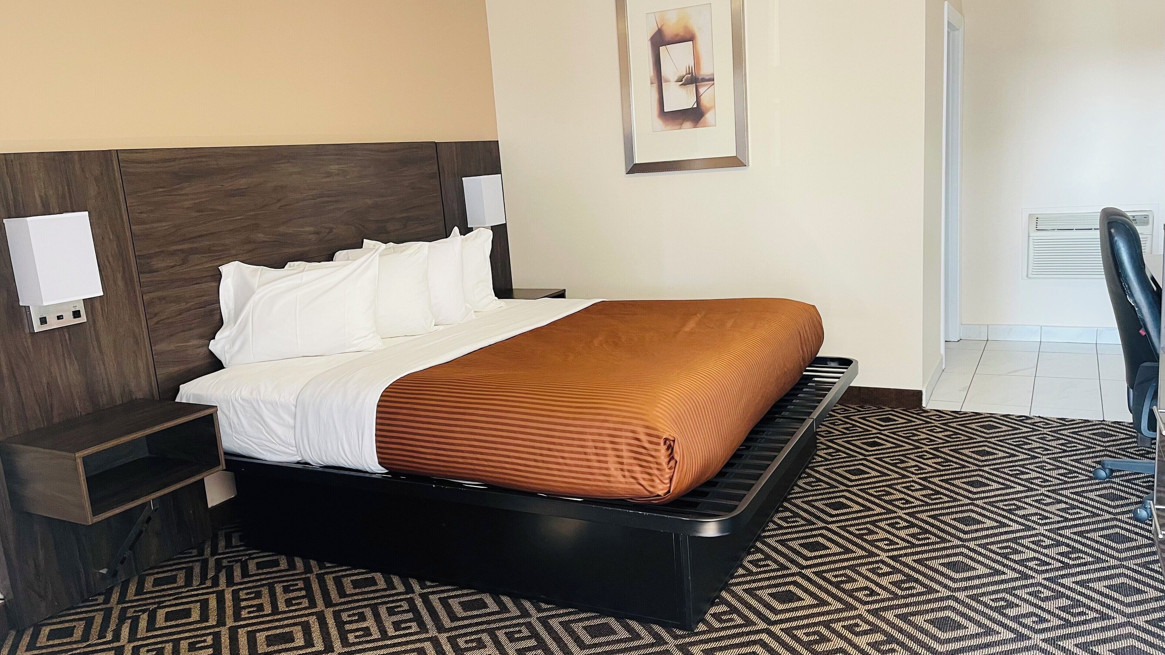 A hotel room with a bed, a nightstand, a wall-mounted light, a framed picture, and a patterned carpet.