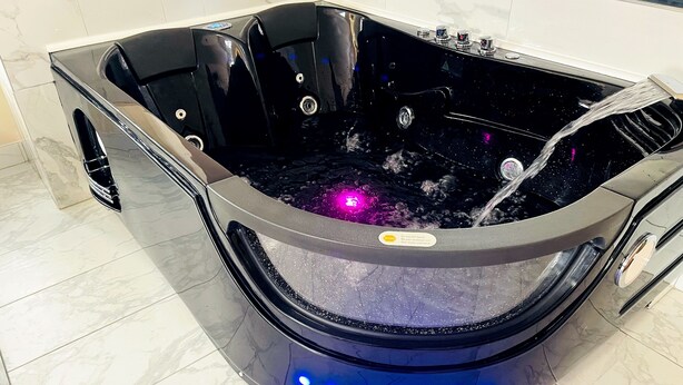 A black hot tub with a control panel and a water pump, surrounded by a tiled floor. - Falls Lodge and Suites (Niagara Falls)