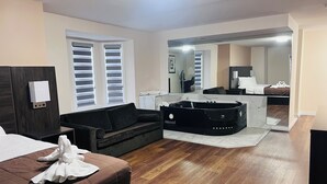 A modern hotel room with a wooden floor, a black sofa, a bed with a brown headboard, and a large mirror reflecting the room. - Falls Lodge and Suites (Niagara Falls)