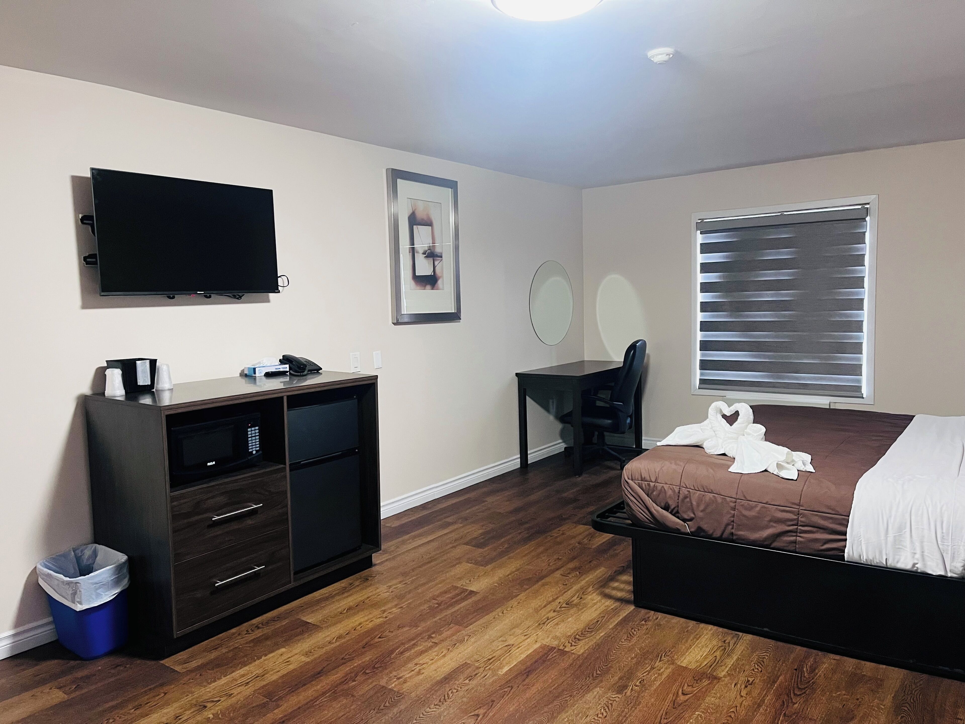 A bedroom with a bed, a desk, a chair, a TV, and a window with blinds.