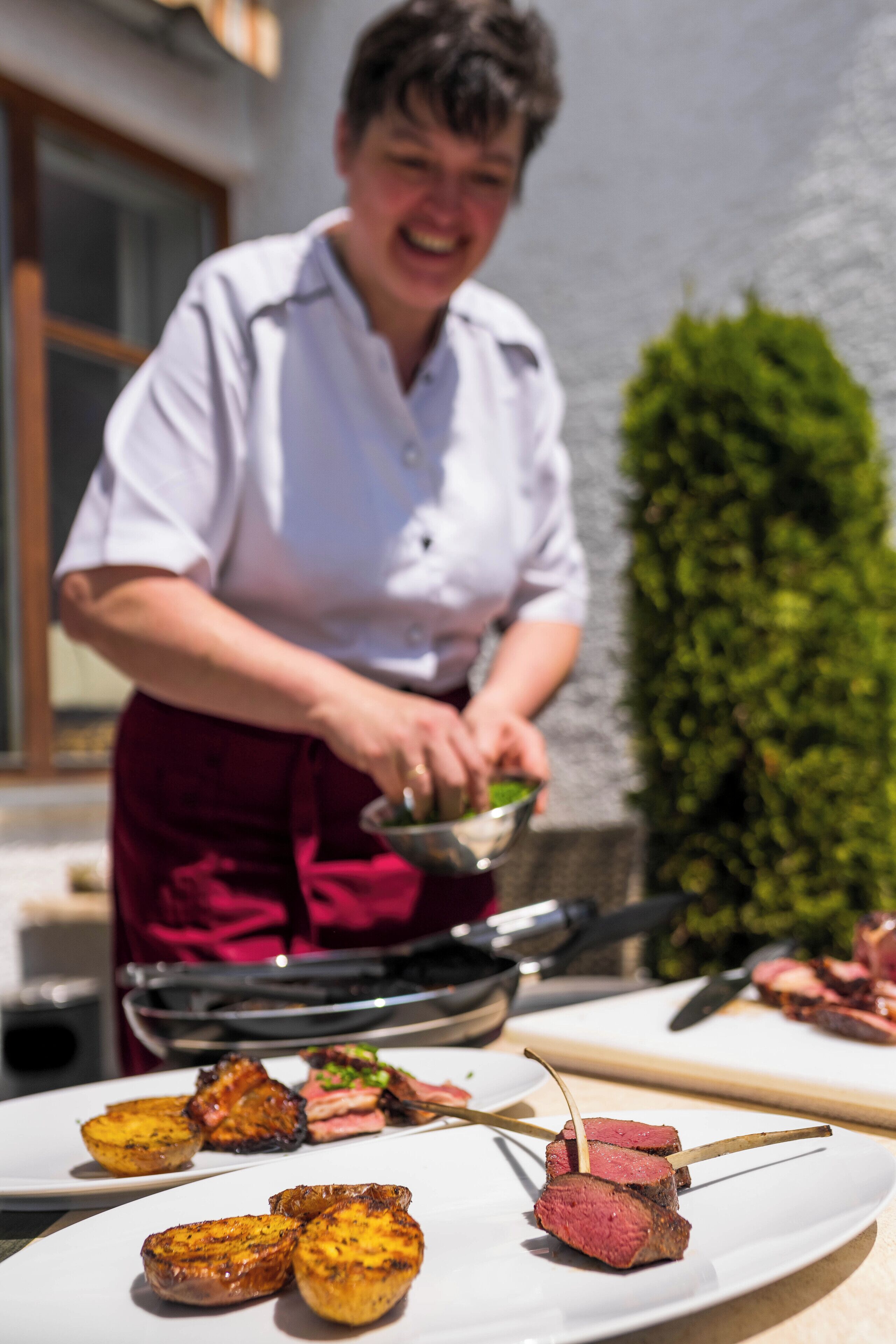 Ein Koch bereitet im Freien Essen zu, er trägt ein weißes Hemd und eine rote Schürze. Im Hintergrund steht ein Grill, auf einem Teller liegen Spieße mit Fleisch und Bratkartoffeln.