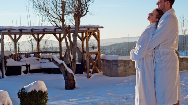 A couple embracing in a snowy outdoor setting with a wooden pergola and snow-covered trees. - Panoramahotel Grobauer (Spiegelau)